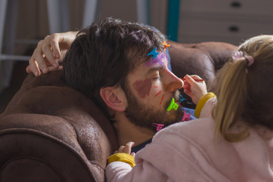 Young father sleeping on the couch while his little daughter paints his face with colorful watercolors