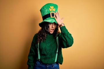 Beautiful curly hair woman wearing green hat with clover celebrating saint patricks day surprised with hand on head for mistake, remember error. Forgot, bad memory concept.