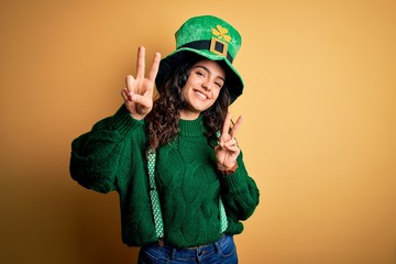 Beautiful curly hair woman wearing green hat with clover celebrating saint patricks day smiling looking to the camera showing fingers doing victory sign. Number two.