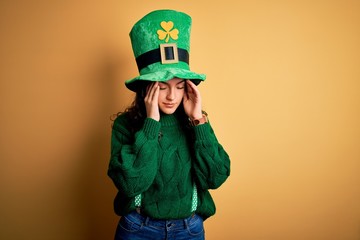 Beautiful curly hair woman wearing green hat with clover celebrating saint patricks day with hand on head for pain in head because stress. Suffering migraine.