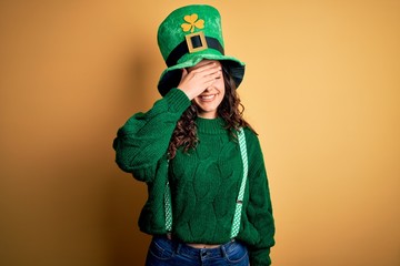 Beautiful curly hair woman wearing green hat with clover celebrating saint patricks day smiling and laughing with hand on face covering eyes for surprise. Blind concept.