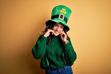 Beautiful curly hair woman wearing green hat with clover celebrating saint patricks day covering ears with fingers with annoyed expression for the noise of loud music. Deaf concept.