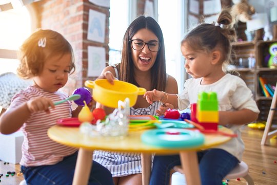 Young beautiful teacher and toddlers playing meals using plastic food and cutlery toy at kindergarten