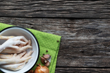 Raw chicken legs lie in a bowl on a wooden table. Near spices and onions with garlic. Green kitchen napkin under the plate. Useful organic product for diets and healthy eating.