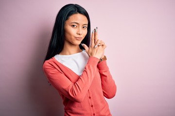 Young beautiful chinese woman wearing casual sweater over isolated pink background Holding symbolic gun with hand gesture, playing killing shooting weapons, angry face