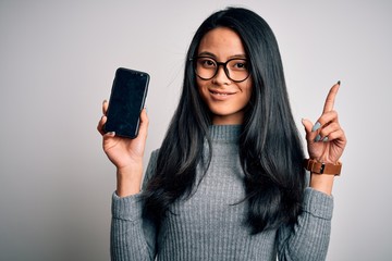 Young beautiful chinese woman holding smartphone over isolated white background surprised with an...