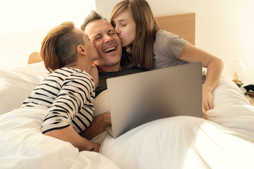 Fototapeta premium Happy relaxed family of using laptop in bed at home. Mom, father and daughter spending time together in the morning