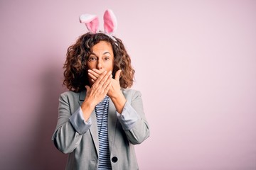 Middle age beautiful woman wearing bunny ears standing over isolated pink background shocked covering mouth with hands for mistake. Secret concept.