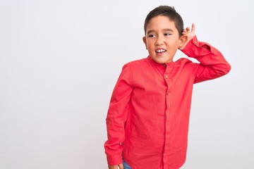 Beautiful kid boy wearing elegant red shirt standing over isolated white background smiling with hand over ear listening an hearing to rumor or gossip. Deafness concept.