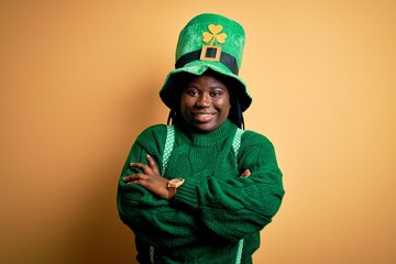 Plus size african american woman with braids wearing green hat with clover on st patricks day happy face smiling with crossed arms looking at the camera. Positive person.