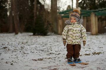 Little boy skier in bright clothes standing on small ski on snow. Background of snowy winter park