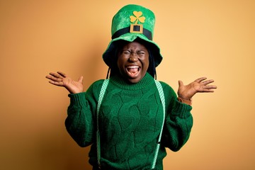 Plus size african american woman with braids wearing green hat with clover on st patricks day celebrating mad and crazy for success with arms raised and closed eyes screaming excited. Winner concept