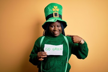 Plus size african american woman wearing green hat holding paper on saint patricks day with surprise face pointing finger to himself