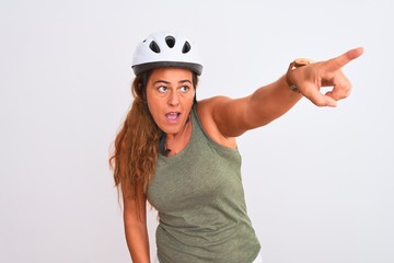 Middle age mature cyclist woman wearing safety helmet over isolated background Pointing with finger surprised ahead, open mouth amazed expression, something on the front