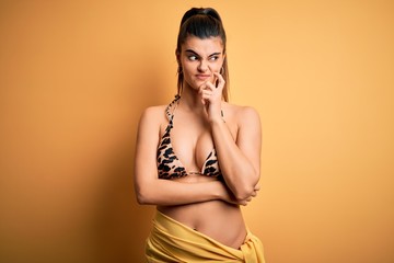 Young beautiful brunette woman on vacation wearing swimwear bikini over yellow background with hand on chin thinking about question, pensive expression. Smiling with thoughtful face. Doubt concept.