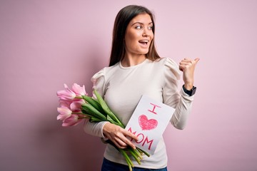 Young beautiful woman with blue eyes holding love mom message and tulips on mothers day pointing and showing with thumb up to the side with happy face smiling