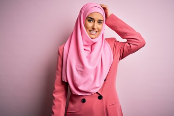 Young beautiful brunette businesswoman wearing pink muslim hijab and business jacket confuse and wonder about question. Uncertain with doubt, thinking with hand on head. Pensive concept.