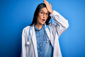 Young beautiful doctor woman wearing stethoscope and glasses over blue background surprised with hand on head for mistake, remember error. Forgot, bad memory concept.