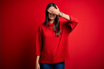 Young beautiful brunette woman wearing casual sweater over isolated red background covering eyes with hand, looking serious and sad. Sightless, hiding and rejection concept