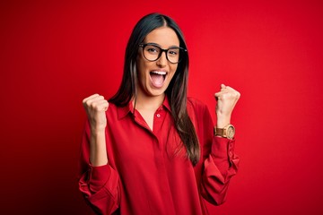 Young beautiful brunette woman wearing casual shirt and glasses over red background celebrating surprised and amazed for success with arms raised and open eyes. Winner concept.