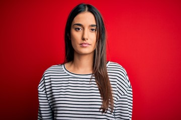 Young beautiful brunette woman wearing casual striped t-shirt over red background Relaxed with serious expression on face. Simple and natural looking at the camera.