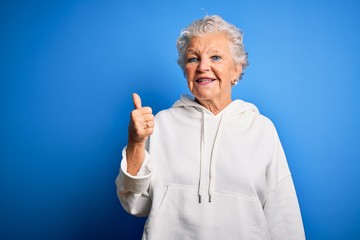 Senior beautiful sporty woman wearing white sweatshirt over isolated blue background doing happy thumbs up gesture with hand. Approving expression looking at the camera showing success.