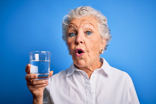 Senior beautiful woman drinking glass of water standing over isolated blue background scared in shock with a surprise face, afraid and excited with fear expression