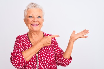 Senior grey-haired woman wearing red casual jacket standing over isolated white background amazed and smiling to the camera while presenting with hand and pointing with finger.