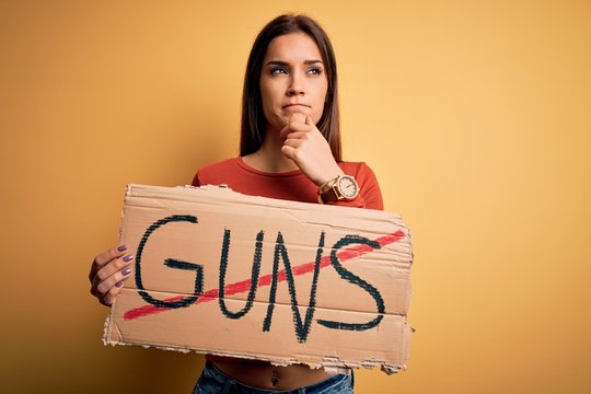 Young beautiful activist woman asking for peace holding banner with stop guns message serious face thinking about question, very confused idea