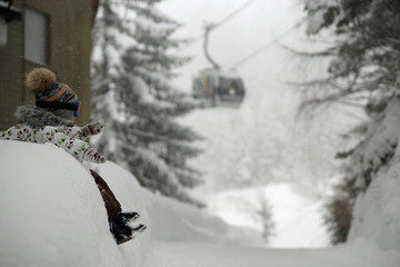 Naklejka premium Small child in ski mask sitting on big snowbank under snowfall. Background of snowy trees, ski cabin going up on cabel