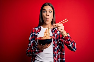 Young beautiful brunette woman eating noodles using chopsticks over red background scared in shock...