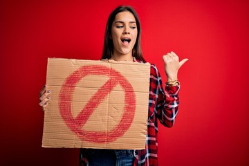 Young beautiful brunette woman holding banner with prohibited signal over red background pointing...