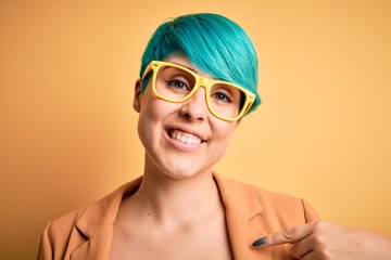 Young beautiful woman with blue fashion hair wearing casual glasses over yellow background with surprise face pointing finger to himself