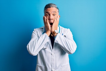 Middle age handsome grey-haired doctor man wearing coat and blue stethoscope afraid and shocked, surprise and amazed expression with hands on face
