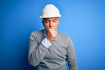 Middle age handsome grey-haired engineer man wearing safety helmet over blue background feeling unwell and coughing as symptom for cold or bronchitis. Health care concept.