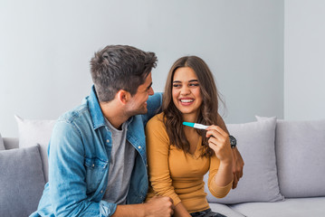 Naklejka na ściany i meble Joyful couple finding out results of a pregnancy test at home. Happy couple looking at pregnancy test. Woman surprising her husband with positive pregnancy test, he seems reasonably pleased