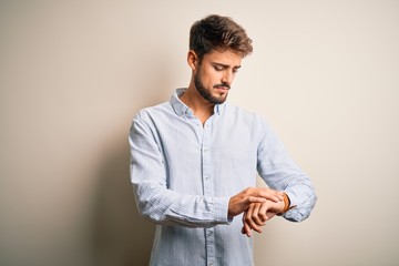 Young handsome man with beard wearing striped shirt standing over white background Checking the time on wrist watch, relaxed and confident