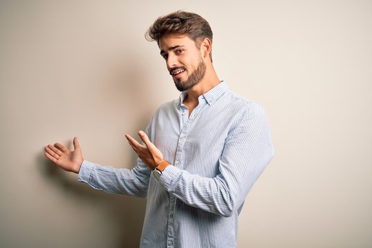 Young handsome man with beard wearing striped shirt standing over white background Inviting to enter smiling natural with open hand