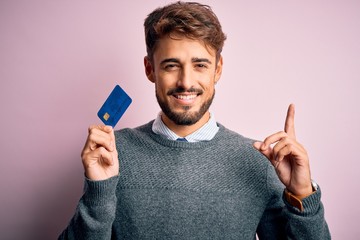 Young customer man with beard holding credit card for payment over pink background surprised with...