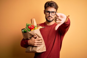 Young man wearing glasses holding paper bag with food over isolated yellow background with angry face, negative sign showing dislike with thumbs down, rejection concept