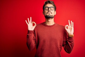 Naklejka premium Young handsome man with beard wearing glasses and sweater standing over red background relaxed and smiling with eyes closed doing meditation gesture with fingers. Yoga concept.
