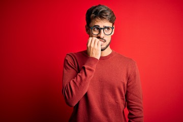 Young handsome man with beard wearing glasses and sweater standing over red background looking stressed and nervous with hands on mouth biting nails. Anxiety problem.