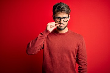 Young handsome man with beard wearing glasses and sweater standing over red background mouth and...