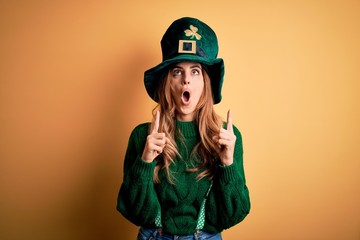 Beautiful brunette woman wearing green hat with clover celebrating saint patricks day amazed and surprised looking up and pointing with fingers and raised arms.