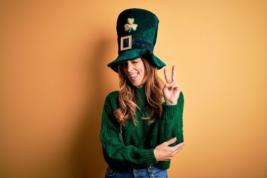 Beautiful brunette woman wearing green hat with clover celebrating saint patricks day smiling with happy face winking at the camera doing victory sign. Number two.