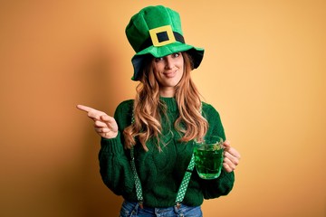 Beautiful woman wearing hat drinking jar of green beverage celebrating saint patricks day very happy pointing with hand and finger to the side