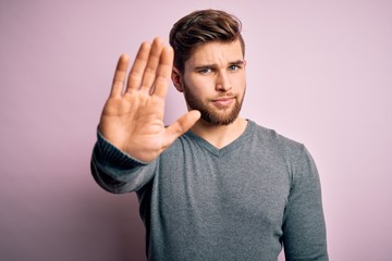 Young handsome blond man with beard and blue eyes wearing casual sweater doing stop sing with palm...