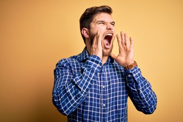 Young blond businessman with beard and blue eyes wearing shirt over yellow background Shouting angry out loud with hands over mouth