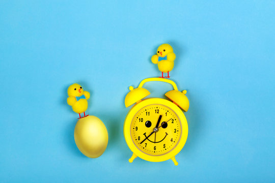 Retro yellow alarm clock with drawn smiling face and toys chicken and colored egg on blue background. Easter concept.