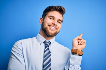 Young blond businessman with beard and blue eyes wearing elegant shirt and tie standing with a big smile on face, pointing with hand and finger to the side looking at the camera.